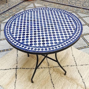 May include: A round mosaic tile table with a blue and white geometric pattern. The table has a black metal frame and legs. The table is sitting on a cream-colored rug.