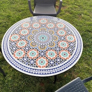 Op de afbeelding: Een ronde mozaïektafel met een kleurrijk geometrisch patroon in blauw, groen, oranje en wit. De tafel is omringd door drie zwarte stoelen op een grasveld. De diameter van de tafel is ongeveer 100 cm.