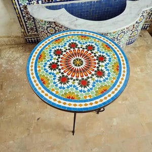 May include: A round mosaic tile table with a black metal base. The tabletop features a colorful geometric pattern with red, orange, green, and blue tiles. The table is approximately 61 cm in diameter.