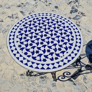 May include: Circular outdoor table with a mosaic tile top. The tabletop has a white background and a repeating pattern of blue stars and geometric shapes. The table has a wrought iron base.