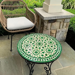 May include: A round outdoor mosaic tile table with a green and white geometric pattern. The table has a black wrought iron base with decorative scrollwork. A wicker chair with a white cushion is next to the table.
