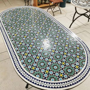 May include: An oval mosaic tile table with a detailed floral pattern in shades of green, blue, yellow, and brown. The table has a white border with a geometric design and black metal legs. The table is a decorative piece.