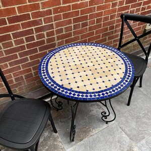 May include: A round bistro table with a mosaic tile top. The table has a black wrought iron base and is flanked by two black chairs. The tile design features a pattern of cream and blue squares and diamonds.