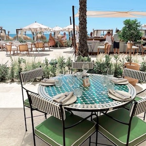 May include: A round outdoor dining table with a teal and white mosaic tile top, set with plates, glasses, and silverware. The table is surrounded by black metal chairs with green cushions and black and white striped backs. The scene is set on a patio near the beach.