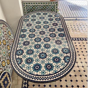 May include: An oval mosaic tile table with a dark blue border. The tabletop features a repeating pattern of blue and white star-shaped designs. The table is surrounded by patterned tile flooring.