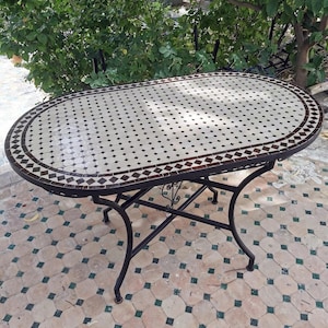 Peut inclure: Table de jardin ovale avec un dessus en mosaïque. Le plateau présente un motif de carreaux blancs avec de petits accents de diamants noirs et une bordure marron. La table a un cadre et des pieds en métal noir.