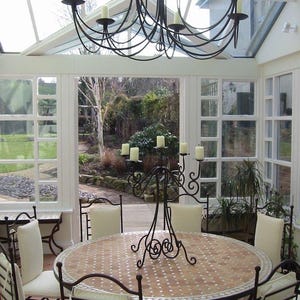 May include: A dining room set with a round table and eight chairs. The table has a mosaic tile top and is surrounded by wrought iron chairs with cream-colored cushions. A wrought iron candelabra and chandelier with candles are also present.