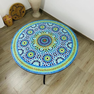 May include: Round mosaic tile table with a black metal base. The tabletop features a vibrant pattern of blue, green, and yellow tiles arranged in a floral design. The table is approximately 30 inches in diameter.
