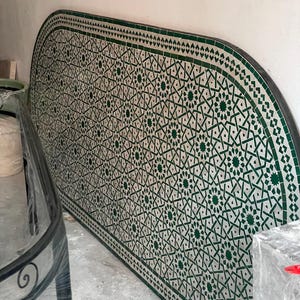 May include: A large, semi-circular table top with a mosaic tile design. The tiles are white with a green geometric pattern. The border is a dark green with a repeating triangle design. The table is leaning against a white wall.