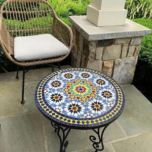 May include: A round mosaic tile table with a black wrought iron base. The tabletop features a colorful geometric pattern with blue, yellow, red, and white tiles. A wicker chair with a white cushion is next to the table.