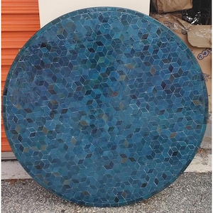 Op de afbeelding: Een ronde tafelblad met een mozaïekontwerp. Het mozaïek bestaat uit kleine, kubusvormige tegels in tinten blauw en blauwgroen. Het tafelblad heeft een donkere rand en staat op een betonnen oppervlak.