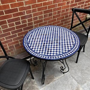 May include: A round outdoor table with a mosaic tile top in blue and white geometric patterns. The table has a black metal frame and is paired with two black chairs. The background features a red brick wall.