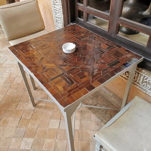 May include: A square table with a mosaic tile top in shades of brown and tan. The table has a light grey metal frame and is accompanied by two chairs with beige cushions. A small white ashtray sits on the table.