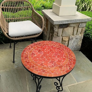 May include: A round mosaic tile table with a red and orange floral pattern sits on a patio. The table has a black metal frame with decorative scrollwork legs. A wicker chair with a white cushion is to the left.