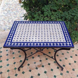 May include: Rectangular outdoor table with a mosaic tile top. The tabletop features a white background with blue diamond and dot patterns, framed by a blue border. The table has a black wrought iron base.