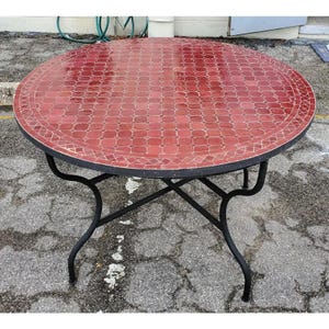 May include: Round outdoor table with a mosaic tile top in shades of red. The table has a black metal frame and legs. The table's diameter is approximately 48 inches.
