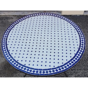 May include: A round outdoor table with a mosaic tile top. The table features a white background with blue diamond and square tile accents. The border is a repeating pattern of blue diamond tiles. The table is supported by a black metal frame.