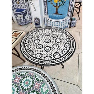 May include: A round mosaic table with a black and white floral pattern. The table has a black border and a dark metal base. Other mosaic pieces are visible in the background.