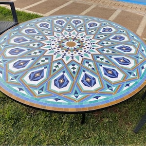 May include: A circular mosaic tile table with a black metal frame. The tabletop has a geometric design in blue, green, white, and gold. The table is set on grass, with two black chairs nearby. The table is approximately 120 cm in diameter.