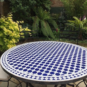 May include: A round outdoor table with a blue and white mosaic tile top. The table is surrounded by black metal chairs with cream-colored cushions. The background features lush greenery and a wooden fence.