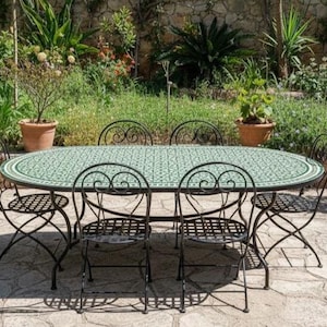 May include: An outdoor dining set featuring an oval table with a green mosaic top and black wrought iron legs. Six matching chairs with curved backs and patterned seats surround the table. The set is placed on a stone patio, surrounded by greenery.