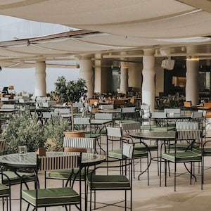 May include: An outdoor restaurant setting with numerous tables and chairs. The chairs have green cushions and striped backs. The tables are round with dark metal frames. Overhead, a beige canopy provides shade, and the floor is light-colored.