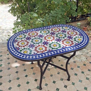 Op de afbeelding: Ovale mozaïektegel tafel met een zwart metalen frame. Het tafelblad heeft een kleurrijk bloemmotief in rood, blauw, geel en groen. De tafel is ongeveer 100 cm lang en 50 cm breed, geschikt voor buitengebruik.