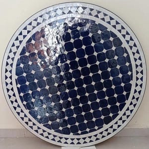 May include: Circular mosaic table with a shiny surface. The tabletop displays a pattern of dark blue octagons, framed by white and black geometric shapes. The table's edge is outlined with a repeating triangle design.