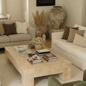 May include: A rectangular coffee table constructed from light beige square tiles with a thick base. The table surface is arranged with several stacks of hardcover books, a small dark bowl, and a ceramic vase filled with dried white flowers.