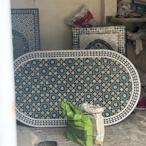 May include: An oval mosaic tile table with a teal, yellow, and black floral pattern. The table is surrounded by other mosaic tile pieces and bags of materials. The table is approximately 183 cm long.