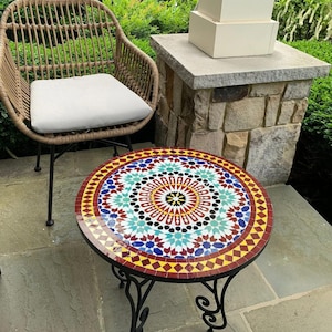May include: Round mosaic tile table with a black wrought iron base. The tabletop features a colorful geometric pattern in red, yellow, blue, and white. A wicker chair with a white cushion is next to the table.