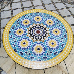 May include: Circular mosaic table featuring a colourful geometric design in blue, yellow, and white. The table has a black edge and is placed on a patterned tile floor and a cream rug.
