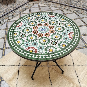 May include: Circular mosaic tile table featuring a green border and complex geometric designs in multiple colours. The table has a black metal base and rests on a cream rug.