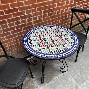 May include: A round mosaic tile table with a black wrought iron base and two black chairs. The table's surface features a pattern of blue, red, green, and white tiles. The chairs have a cross-back design.