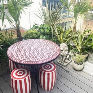 May include: Outdoor patio set with a round mosaic tile table and three matching striped stools. The table has a black metal frame. The stools and table have red and white patterns. The patio is surrounded by lush greenery.