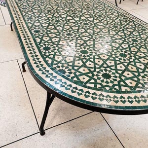 May include: An oval mosaic tile table. The tabletop features a repeating geometric pattern in green and cream. The table has a black metal frame and legs. The table's design is reminiscent of Moroccan style.