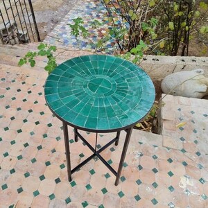 May include: A round outdoor side table with a mosaic tile top in shades of green. The table has a black metal frame and legs. The table's diameter is approximately 61 cm.