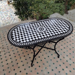 May include: An oval mosaic table with a black and white checkered pattern top. The table has a decorative black metal frame and legs. The table's dimensions are approximately 152 cm long and 76 cm wide.