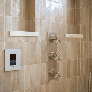 Peut inclure: Mur de douche carrelé beige avec une étagère encastrée et des robinets chromés. La douche comprend un panneau de commande, trois robinets en croix et une douchette.