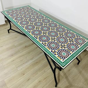 May include: A rectangular table with a mosaic tile top featuring a repeating geometric pattern in shades of blue, brown, and teal. The table has a green border and black metal legs. The table is approximately 152 cm long.