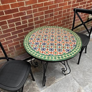 May include: A round outdoor table with a mosaic tile top featuring a geometric pattern in red, blue, and green. The table has a black metal frame and is surrounded by two black chairs. The table's diameter is approximately 30 inches.