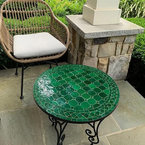 May include: A round outdoor table with a mosaic green tile top and black wrought iron legs. A wicker chair with a white cushion is next to the table. The table is approximately 24 inches in diameter.