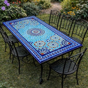 May include: Rectangular outdoor dining table with a mosaic tile top in shades of blue, red, and white. The table is surrounded by six black wrought iron chairs. The table is set on a grassy lawn, with greenery in the background.