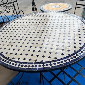 May include: A large, round outdoor table with a mosaic tile top. The table features a white and blue geometric pattern with a dark blue border. The table is supported by a black metal frame. Other tables and chairs are visible in the background.
