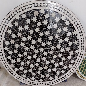 May include: Round mosaic table with a black and white star pattern. The table has a decorative border with a repeating geometric design. The mosaic is made of small tiles, creating a detailed and intricate surface.