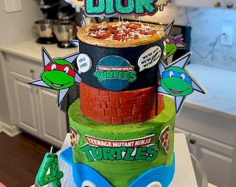 May include: A multi-tiered birthday cake featuring Teenage Mutant Ninja Turtles theme. The cake includes a pizza layer, brick-like design, and a turtle shell base. Toppers read "HAPPY BIRTHDAY" and "DIOR". Includes a number 4 candle.