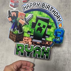 May include: A Minecraft-themed birthday cake topper with the words "Happy Birthday Ryan" in green pixelated letters. The topper features Minecraft characters, TNT blocks, and a number 9 candle.
