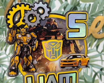 May include: A yellow and black Transformers cake topper with a Bumblebee character, a silver car, and the name "Liam" in silver letters. The topper also features gears and the Transformers logo.