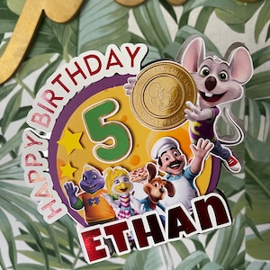 May include: A colorful cake topper featuring Chuck E. Cheese characters, including a purple dinosaur, a yellow bird, a brown dog, and a chef. The topper also includes a gold coin with the words "Where a Kid Can Be a Kid" and "Chuck E. Cheese" and the number 5. The topper says "Happy Birthday Ethan".