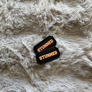 Puede incluir: Dos parches rectangulares negros con la palabra "STUNNED" en letras naranjas. Los parches est&aacute;n sobre una superficie gris y esponjosa.
